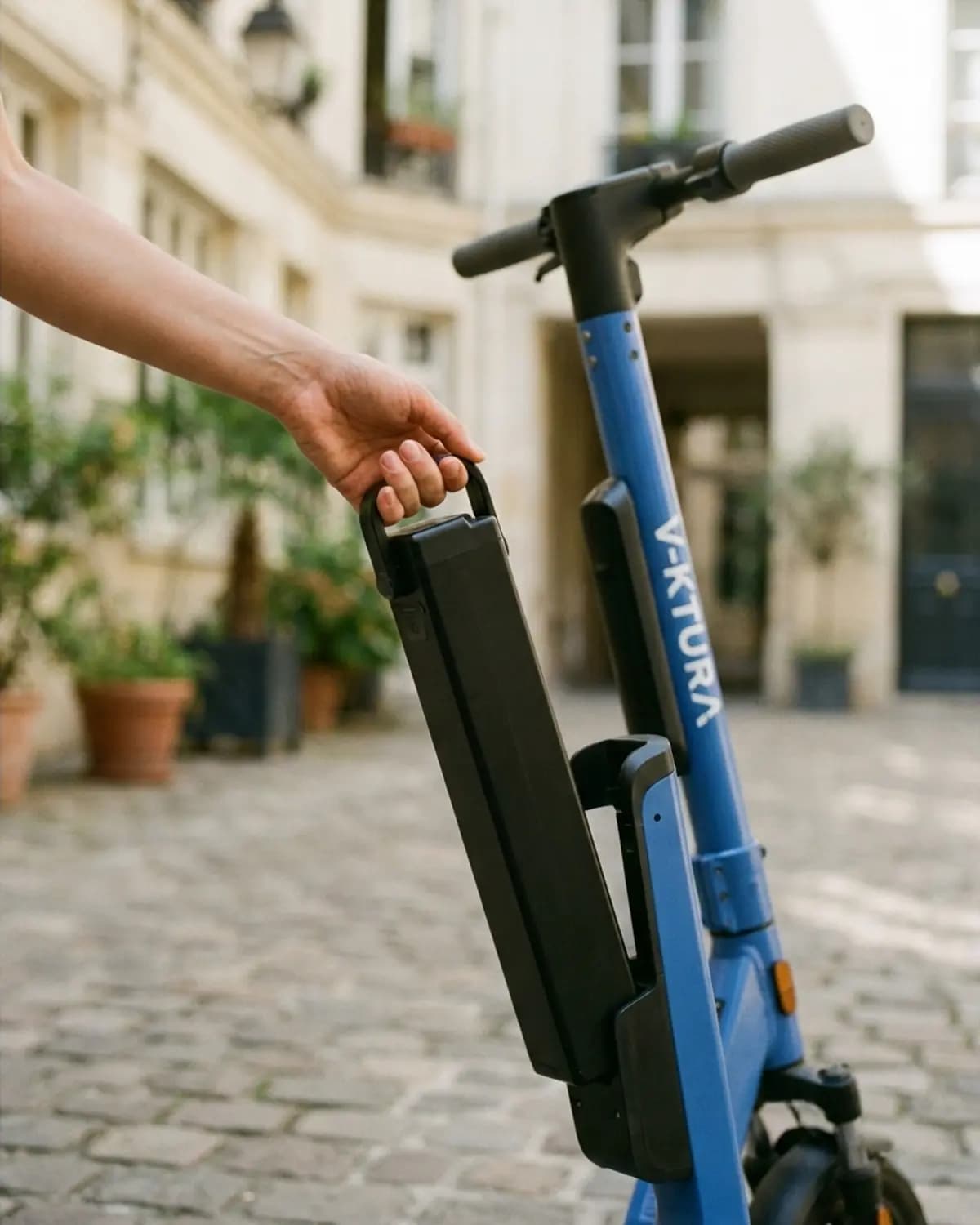 Une main retire la batterie noire fixée au tube bleu d'une trottinette V-KTURA, dans une cour pavée parisienne.